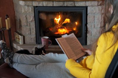 Femme qui lit un livre confortablement installée devant une cheminée avec un feu allumé