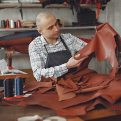 Un artisan examine une pièce de cuir entre ses mains, sur son établi trônent d'autres peaux de mouton